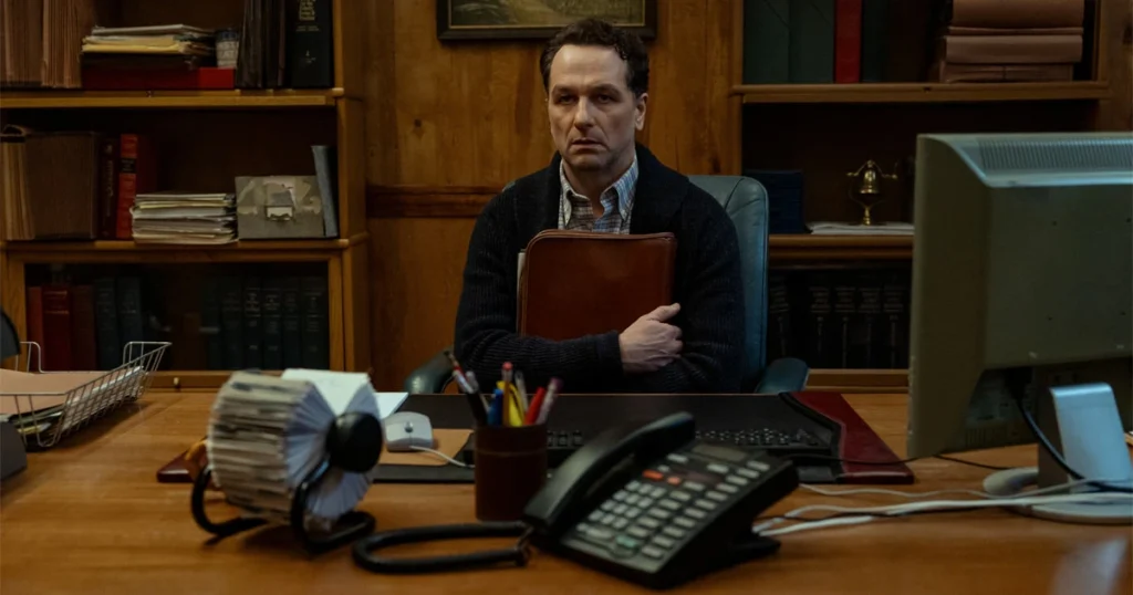 Actor Matthew Rhys as Mayor Tom Loftis sitting anxiously at a wooden office desk in the Apple TV series Widow's Bay.
