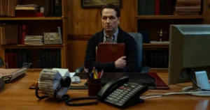 Actor Matthew Rhys as Mayor Tom Loftis sitting anxiously at a wooden office desk in the Apple TV series Widow's Bay.