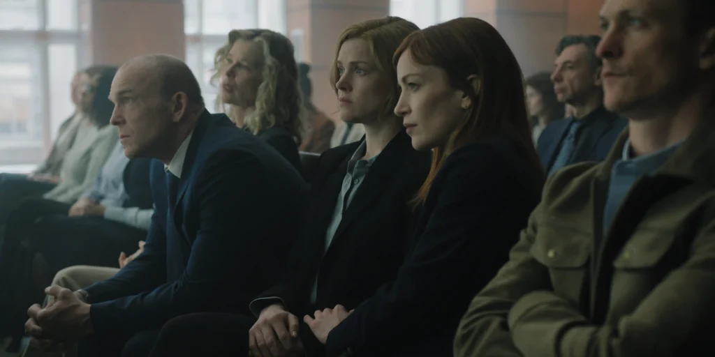 "People sit in rows in a courtroom or formal setting, dressed in business attire, attentively focused on proceedings taking place out of frame, with large windows and neutral lighting in the background."