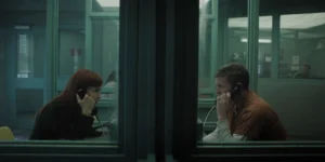 "A woman and a man in prison uniform sit across glass in a visitation room using phones to talk. The setting is dimly lit with institutional green walls and other people are visible in the background."