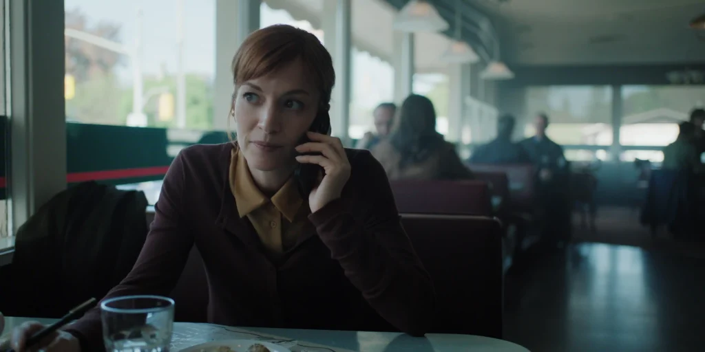 "A woman with red hair sits in a diner booth talking on her phone, with a glass of water and a plate of food on the table; the diner is softly lit with other patrons in the background."