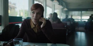 "A woman with red hair sits in a diner booth talking on her phone, with a glass of water and a plate of food on the table; the diner is softly lit with other patrons in the background."