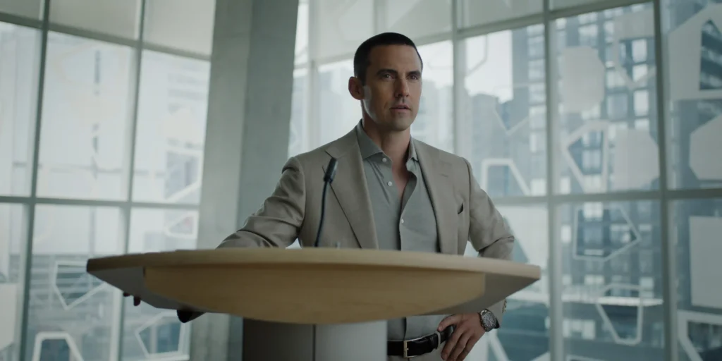 "Man in a suit standing at a podium with a microphone in a modern office or conference room, large windows and cityscape in the background."