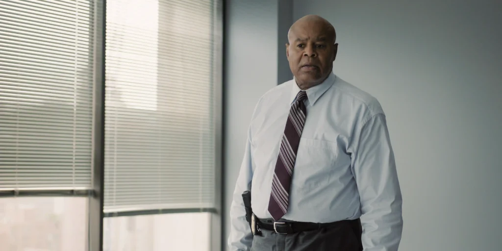 "Man in a dress shirt and tie standing in a modern office with large windows and blinds, neutral expression, daylight coming through the windows."