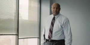 "Man in a dress shirt and tie standing in a modern office with large windows and blinds, neutral expression, daylight coming through the windows."