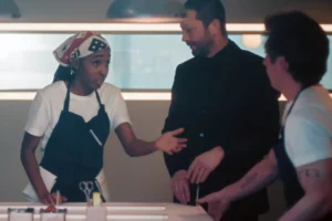 Sydney wearing a chef’s apron and bandana, gesturing during an intense conversation with Richie in a black suit and Carmy in a kitchen apron in The Bear Season 5