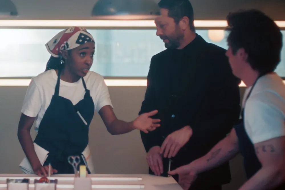 Sydney wearing a chef’s apron and bandana, gesturing during an intense conversation with Richie in a black suit and Carmy in a kitchen apron in The Bear Season 5