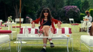 Asha Banks as Noah sitting on an outdoor white couch with her legs crossed, wearing a red jacket, black sunglasses, and a crop top in a scene from Your Fault: London