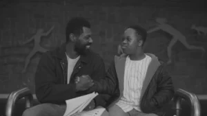 A black and white film still from Color Book showing William Catlett as Lucky and Jeremiah Alexander Daniels as Mason sitting together on a bench
