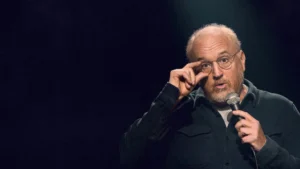 Louis CK holding his glasses while on stage in his Netflix special Ridiculous