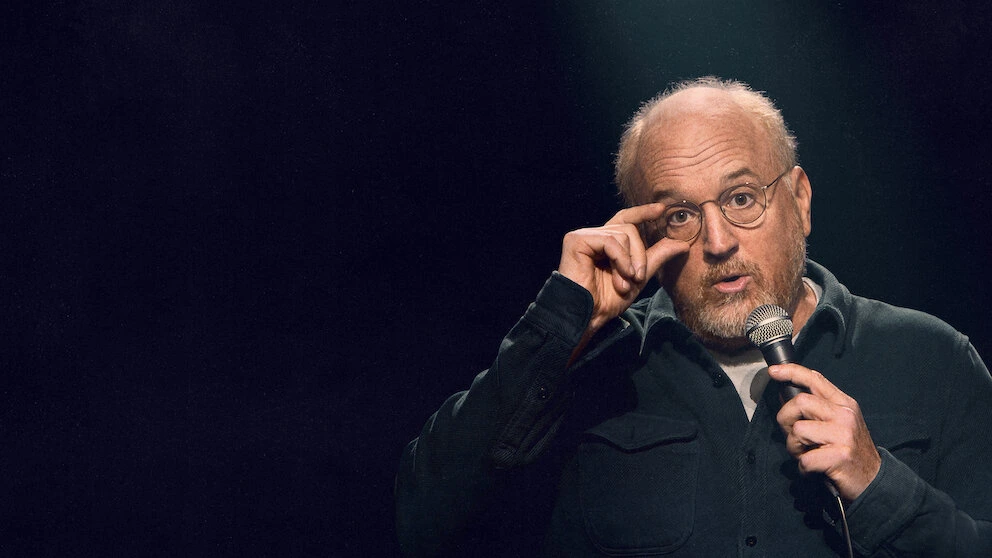 Louis CK holding his glasses while on stage in his Netflix special Ridiculous