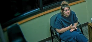 A surveillance camera still of Taylor Parker sitting in an interrogation room chair, wearing a dark short sleeved top, gesturing with her hands while speaking