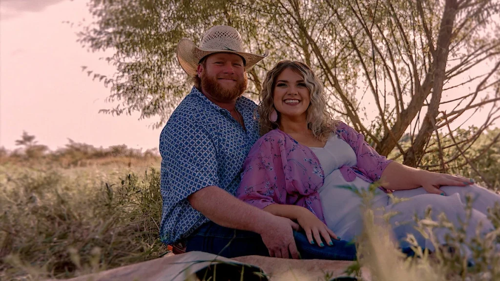 A portrait of Wade Griffin and Taylor Parker, featured in Maternal Instinct on Netflix. Griffin, wearing a cowboy hat and a patterned blue shirt, is seated beside Parker, who wears a light-colored dress and a lavender floral kimono. They are sitting in a grassy area with a large tree in the background