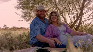 A portrait of Wade Griffin and Taylor Parker, featured in Maternal Instinct on Netflix. Griffin, wearing a cowboy hat and a patterned blue shirt, is seated beside Parker, who wears a light-colored dress and a lavender floral kimono. They are sitting in a grassy area with a large tree in the background