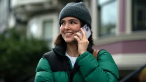 Zoey Deutch as Jill wearing a puff jacket and hat and smiling while using her smart phone in Voicemails for Isabelle