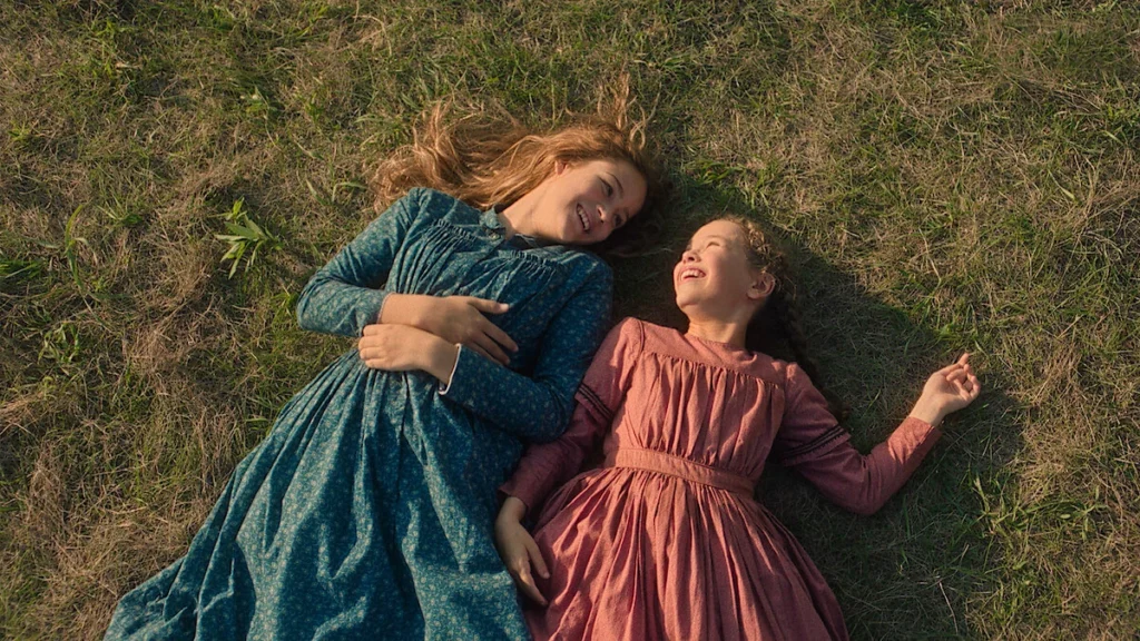 An overhead view of Mary and Laura Ingalls lying down in the green grass, wearing pioneer dresses from Netflix's Little House on the Prairie