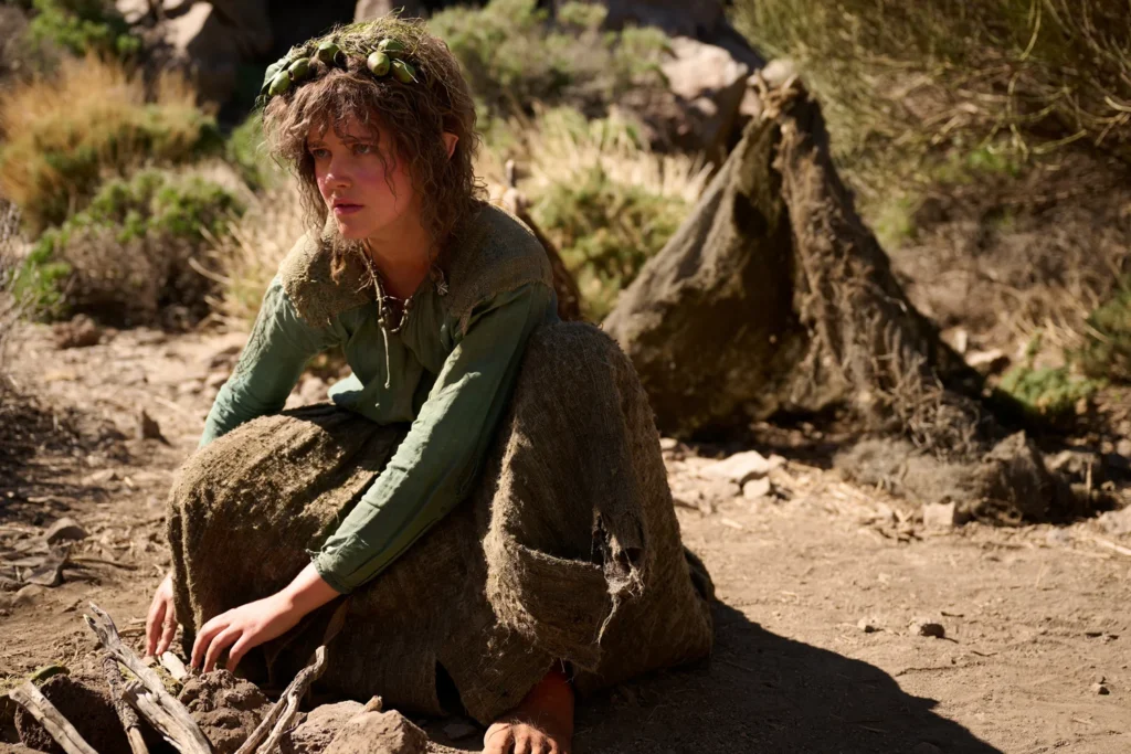 Markella Kavenagh as Nori Brandyfoot crouching in a earthy green dress with twigs woven into her curly hair in The Rings of Power