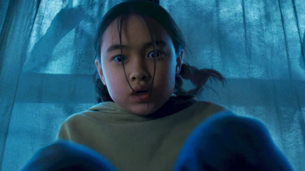 A young girl with pigtails looks down nervously in front of a blue-lit curtain in Netflix's The Last House