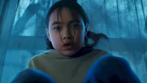 A young girl with pigtails looks down nervously in front of a blue-lit curtain in Netflix's The Last House