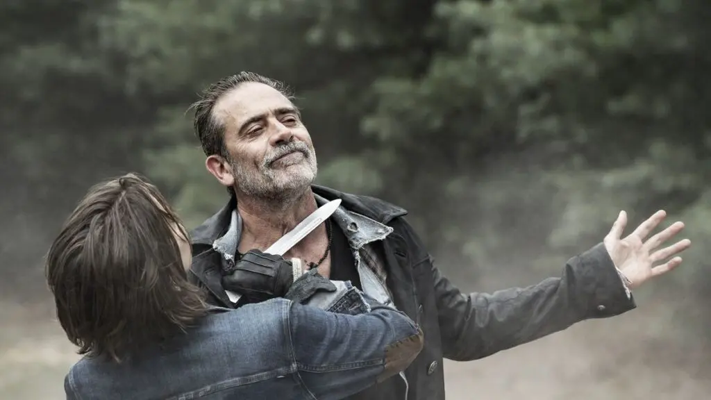 Maggie holding a large knife against Negan's throat while he smirks with arms outstretched in The Walking Dead: Dead City